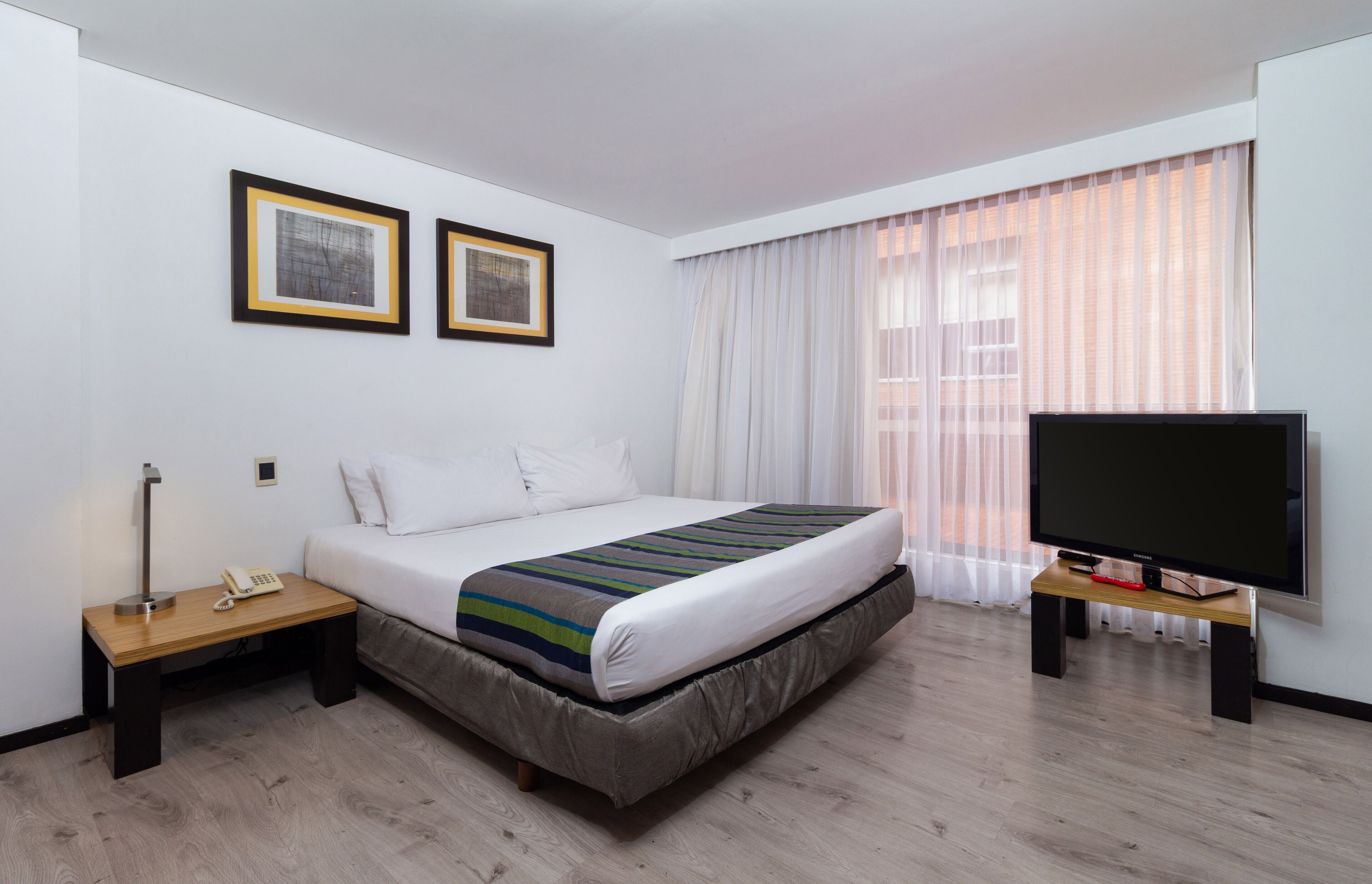 Habitación de hotel con una cama, un televisor sobre un soporte, una mesita de noche con teléfono y dos cuadros en la pared.