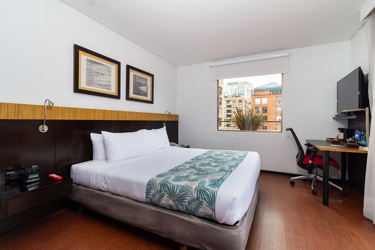 A hotel room with a bed, a desk, a TV, and a window with a view of buildings.