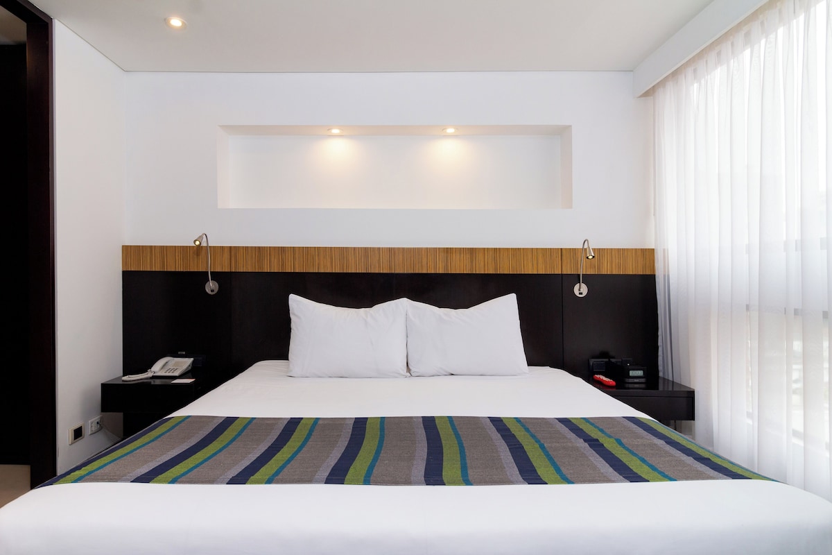 A hotel room with a large bed, a wooden headboard, a bedside table with a phone, and a nightstand with a clock.