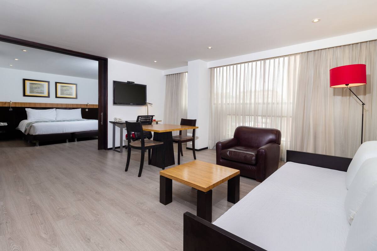 A hotel room with a bed, a desk, a chair, a sofa, a TV, and a lamp.