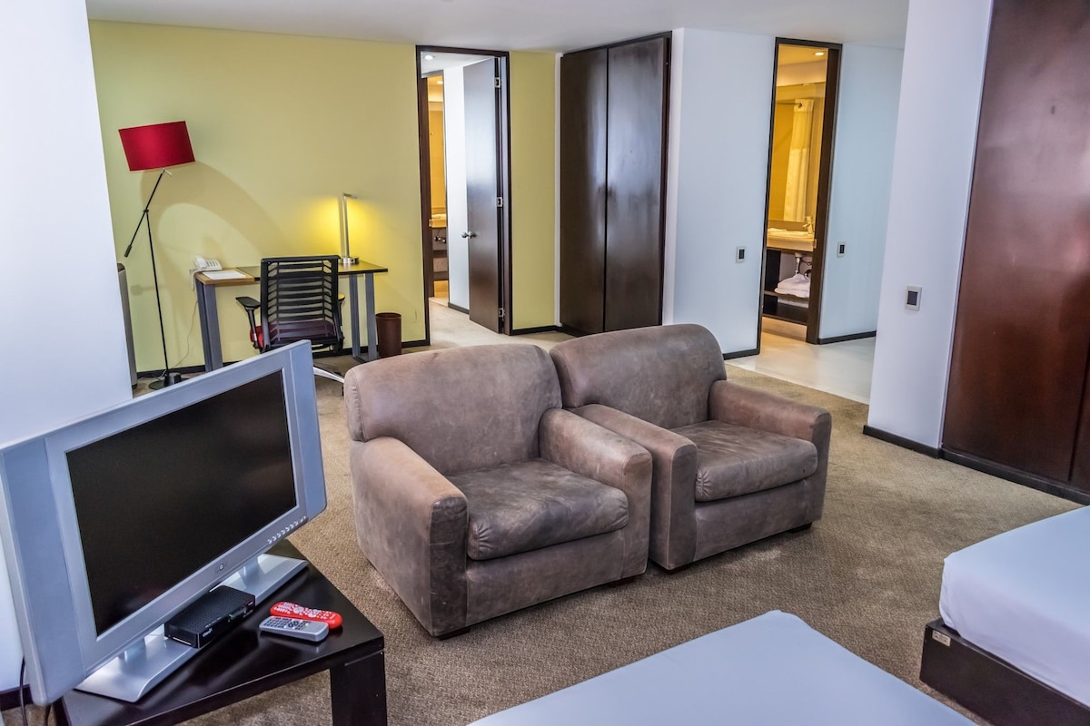A living room with a flat-screen TV, a two-seater sofa, a desk with a lamp, and a chair.