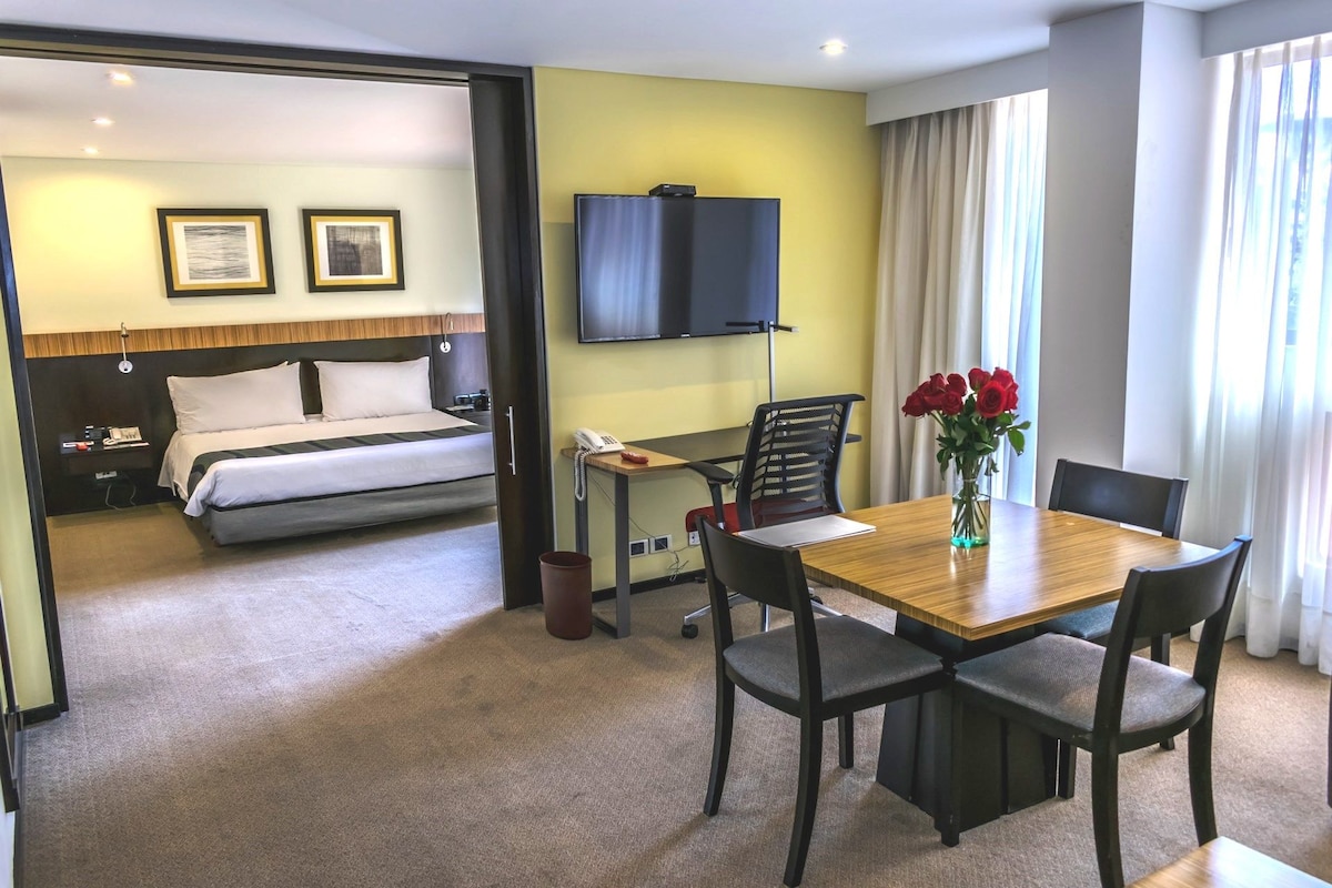 A hotel room with a bed, desk, chair, TV, and a dining table with chairs.