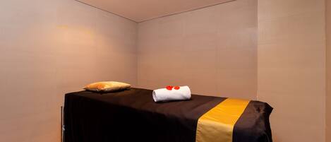 A single bed with a black and gold bedspread, a white pillow, and a rolled towel with red heart-shaped objects on top.