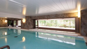 Uma grande piscina interior com água azul cristalina, paredes de pedra e vista para o verde lá fora.