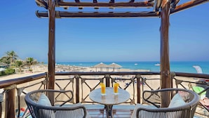 Una terraza de madera con sillas de mimbre y una mesa, con vistas a una playa con sombrillas y cielo despejado.