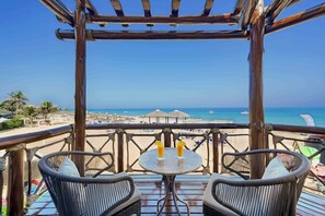 Eine Holzterrasse mit Korbstühlen und einem Tisch mit Blick auf einen Strand mit Sonnenschirmen und blauem Himmel.