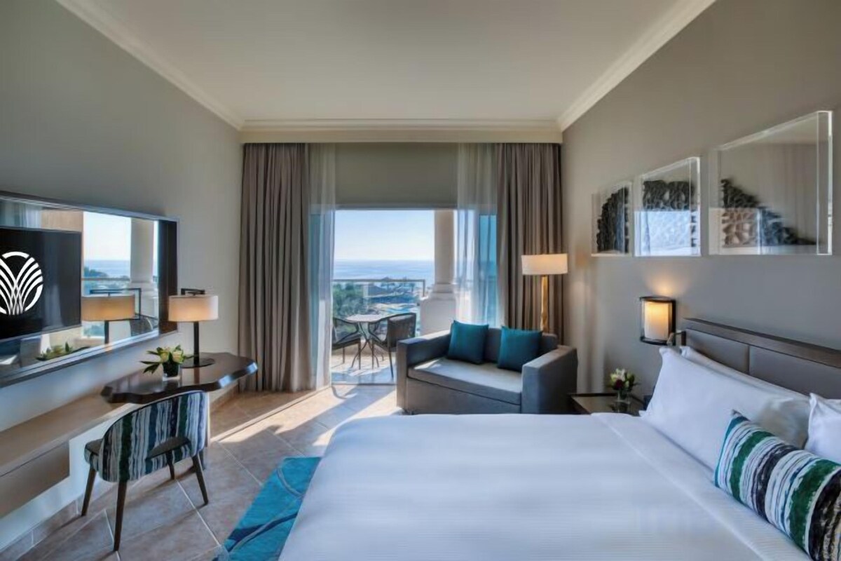 A hotel room with a large bed, a desk with a chair, a television, and a balcony with a view.