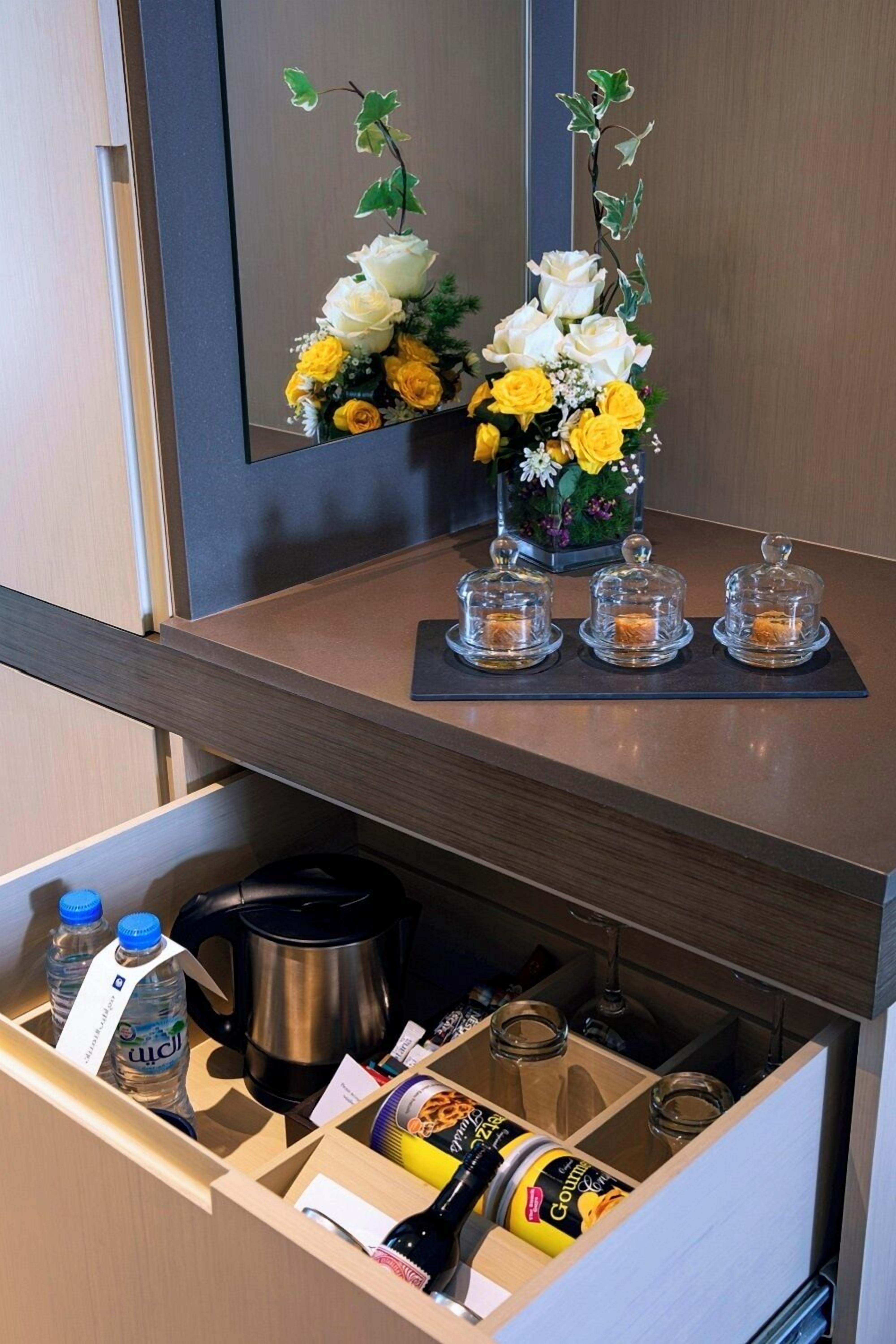 Ein Hotelzimmer mit Schreibtisch aus Holz, einem Spiegel, einer Blumenkomposition und einer Schublade mit Pflegeartikeln und einem Wasserkocher.