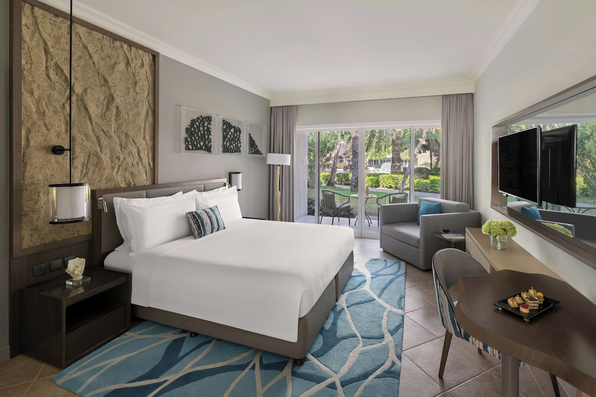 Una habitación de hotel con una cama grande, un escritorio con televisor, un balcón con vistas a palmeras y un comedor con mesa y sillas.