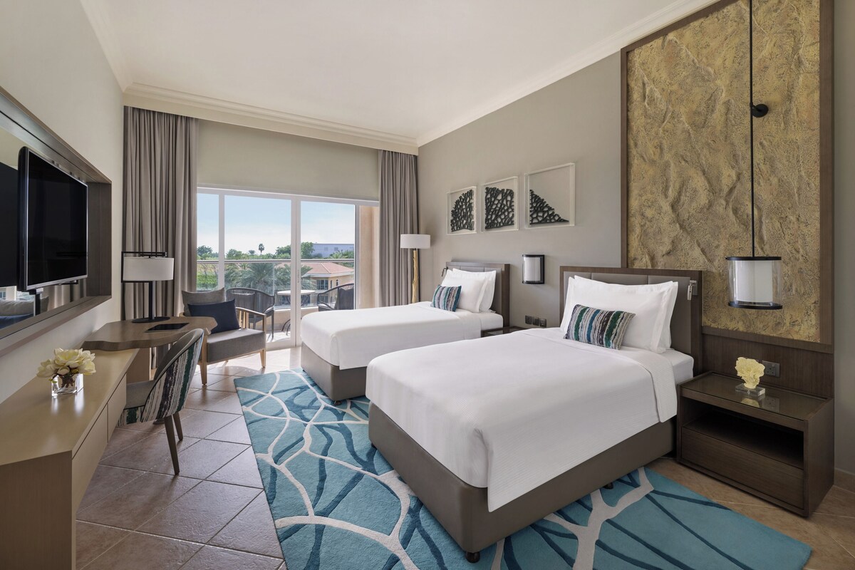 Habitación de hotel con dos camas, un escritorio con televisor, una silla, vistas al balcón y una obra de arte grande en la pared.