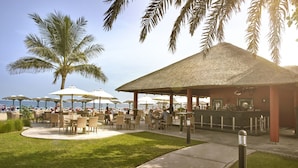 An outdoor seating area with tables and chairs, a bar, and palm trees. - Fujairah Rotana Resort & Spa (Al Aqah)