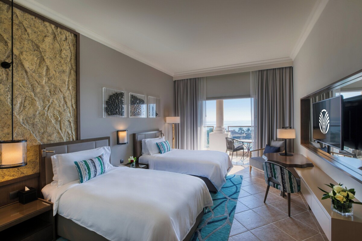 Habitación de hotel con dos camas, televisión, escritorio y balcón con vistas.