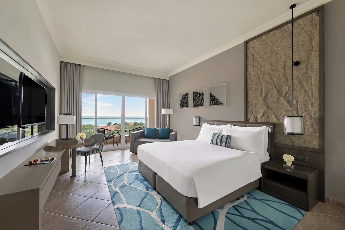 Habitación de hotel con cama, televisión, escritorio, silla, mesita de noche y vista al océano.