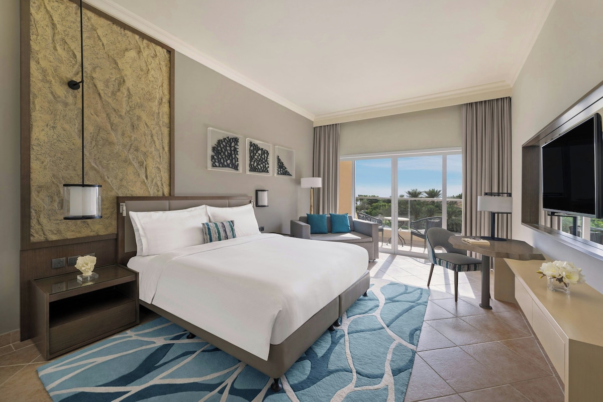 Habitación de hotel con una cama grande, un escritorio, una silla, un televisor y un balcón con vistas.