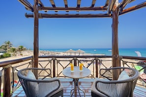 Terraza de madera con sillas de mimbre y una mesa, con vista a una playa que tiene sombrillas y sillas de playa.