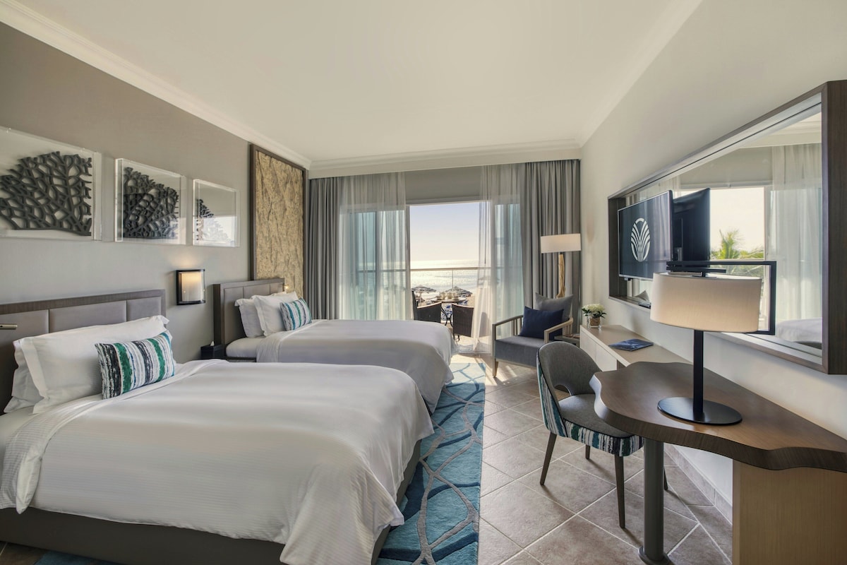 Habitación de hotel con dos camas, un escritorio, una silla, un televisor y vista al mar.