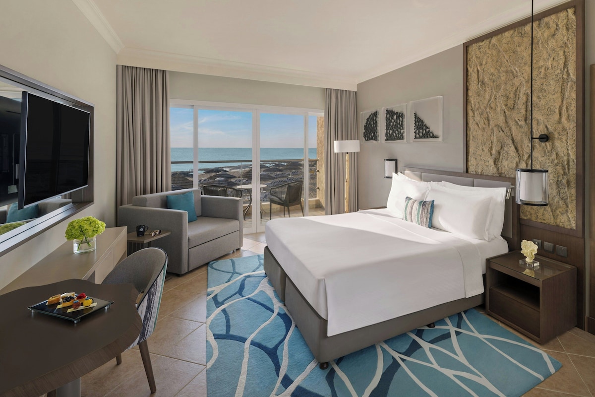 Habitación de hotel con cama, televisor, mesa de comedor y vista a la playa.