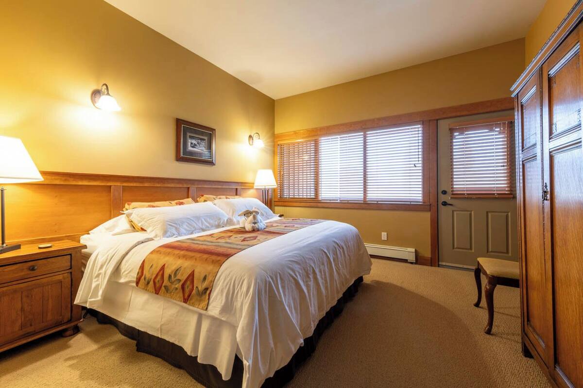 Ein Schlafzimmer mit einem großen Bett, zwei Nachttischlampen, einem gerahmten Bild an der Wand und einer Tür mit Fenster darüber.