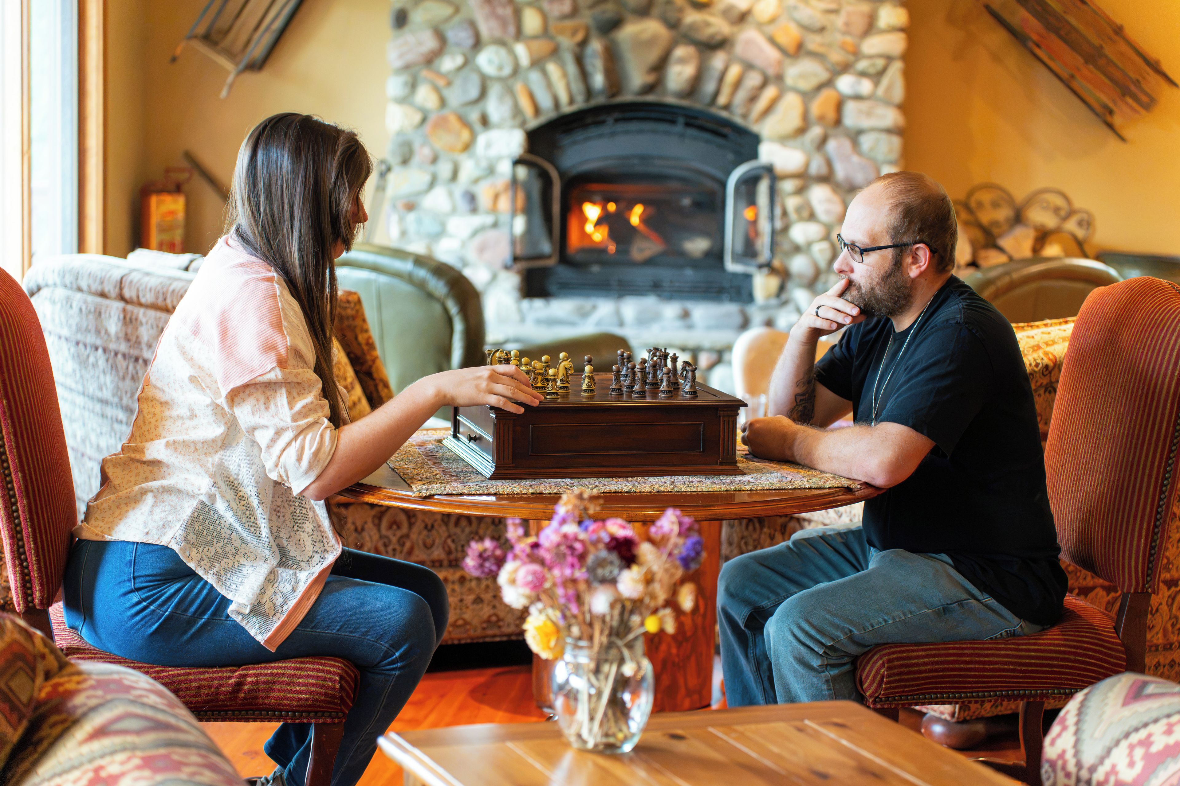 A man and a woman sitting across from each other at a wooden table with a chessboard, in a room with a stone fireplace and patterned furniture.