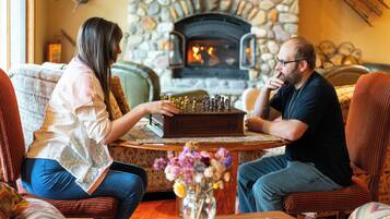 A man and a woman sitting across from each other at a wooden table with a chessboard, in a room with a stone fireplace and patterned furniture.