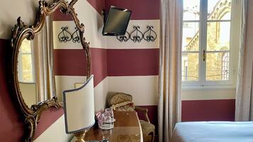 A hotel room with a large ornate mirror, a television mounted on the wall, a bed, a chair, and a table with a lamp and a remote control.