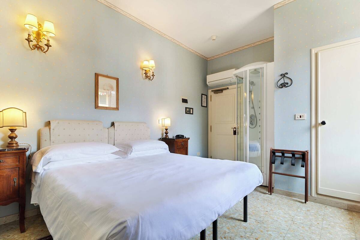A neatly made bed with white linens, a wooden nightstand with a lamp, a wall-mounted light fixture, and a door with a towel rack.