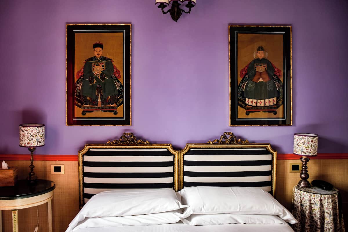 A hotel room with a double bed, two framed paintings on the wall, a chandelier, and a small table with a lamp.