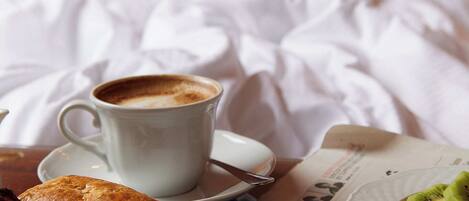 A bed with white linens, a cup of coffee, pastries, and a newspaper on a table.