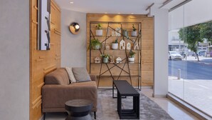 Una sala de estar moderna con un sofá, una mesa redonda de centro y una mesa rectangular negra. Una pared de madera presenta un patrón geométrico y un estante con plantas en macetas y pequeñas decoraciones con forma de casa.