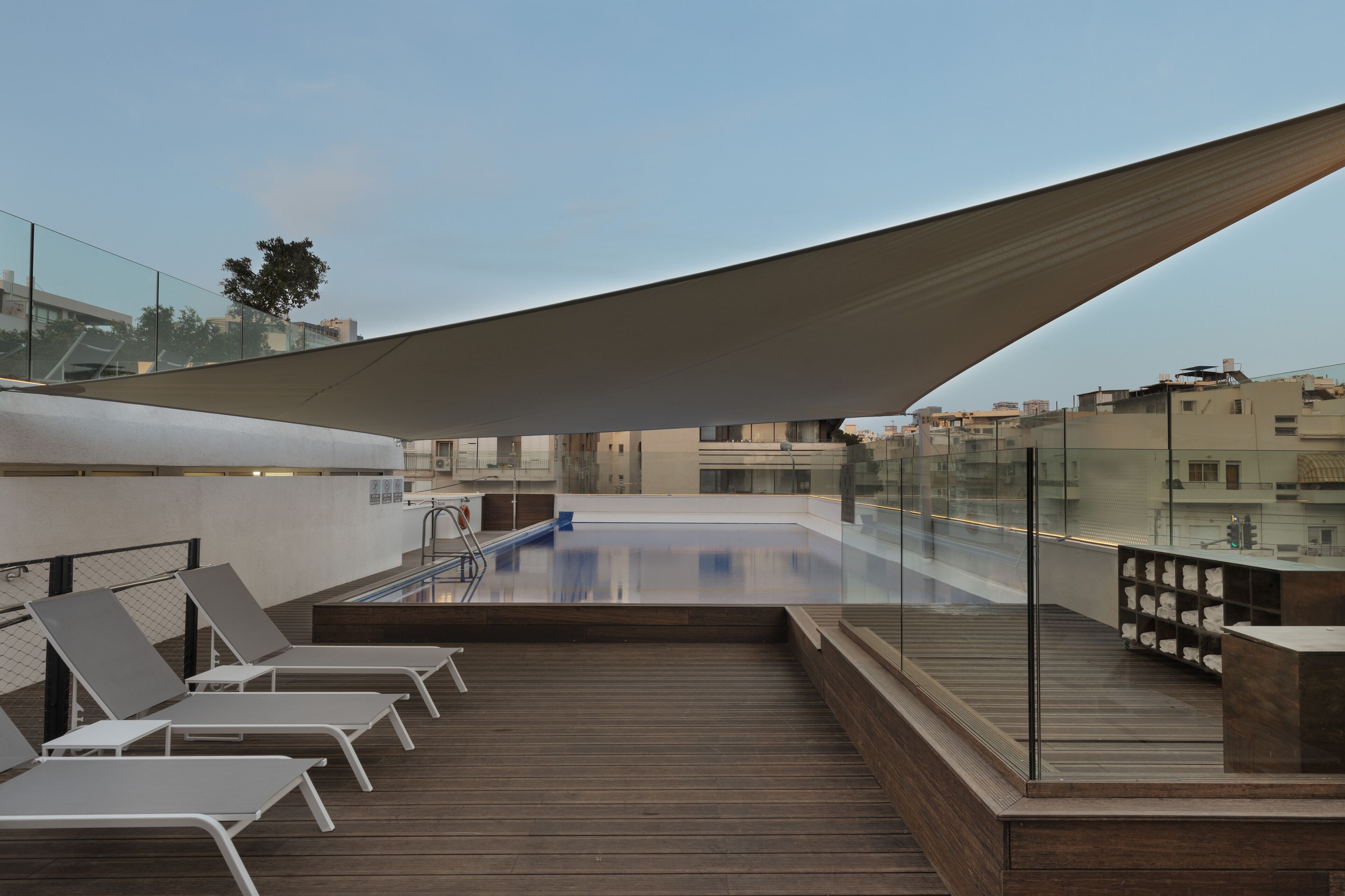 A rooftop pool area with lounge chairs and a glass railing overlooking a cityscape.