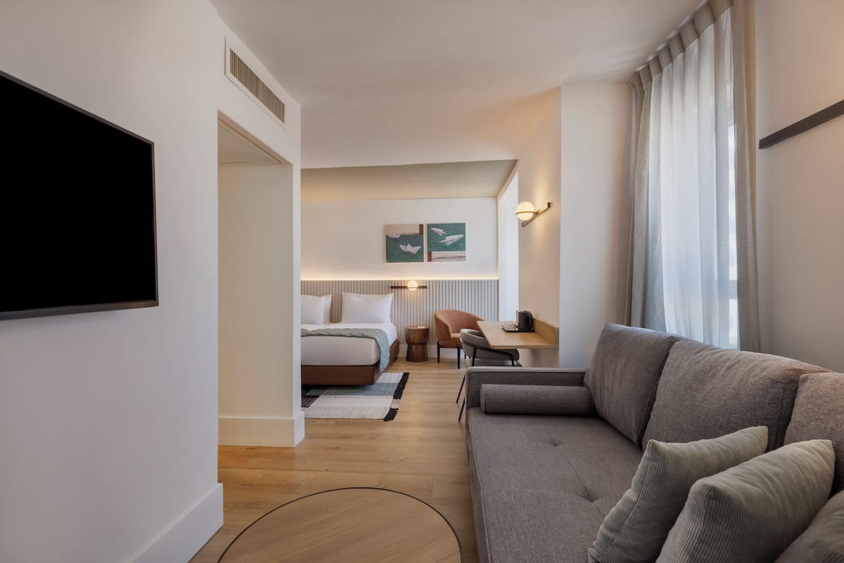 A modern hotel room with a large bed, a sofa, a desk, and a flat-screen TV.