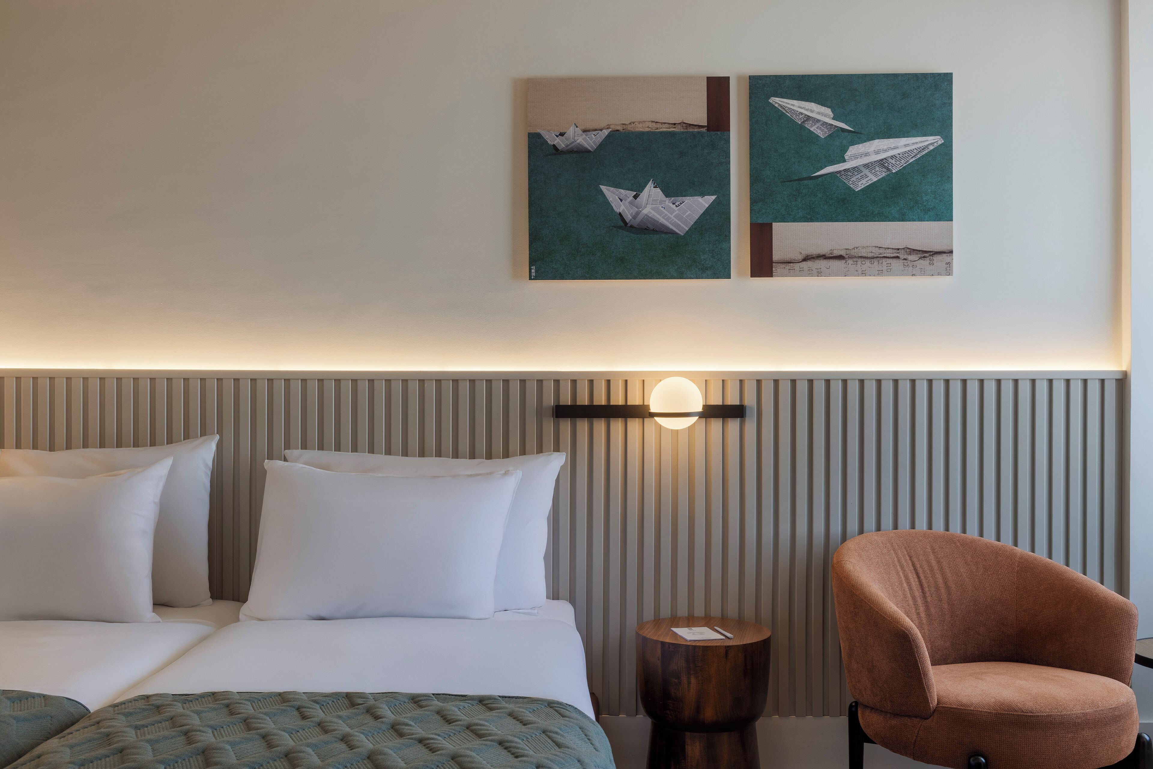 A hotel room with a bed, pillows, a round wooden side table, a chair, and wall art depicting paper airplanes.