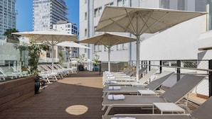 Un área de lounge en la azotea con sillones de sol y sombrillas, rodeada por una terraza de madera y con vistas a un paisaje urbano.