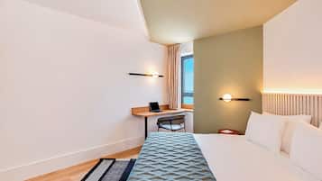 A hotel room with a bed, a desk with a chair, a laptop, a nightstand, and a window with curtains.