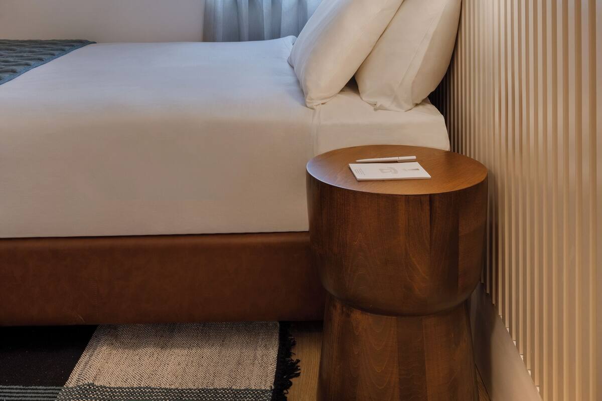 A hotel room with a bed, a round wooden nightstand, a wall-mounted mirror, and a window with sheer curtains.