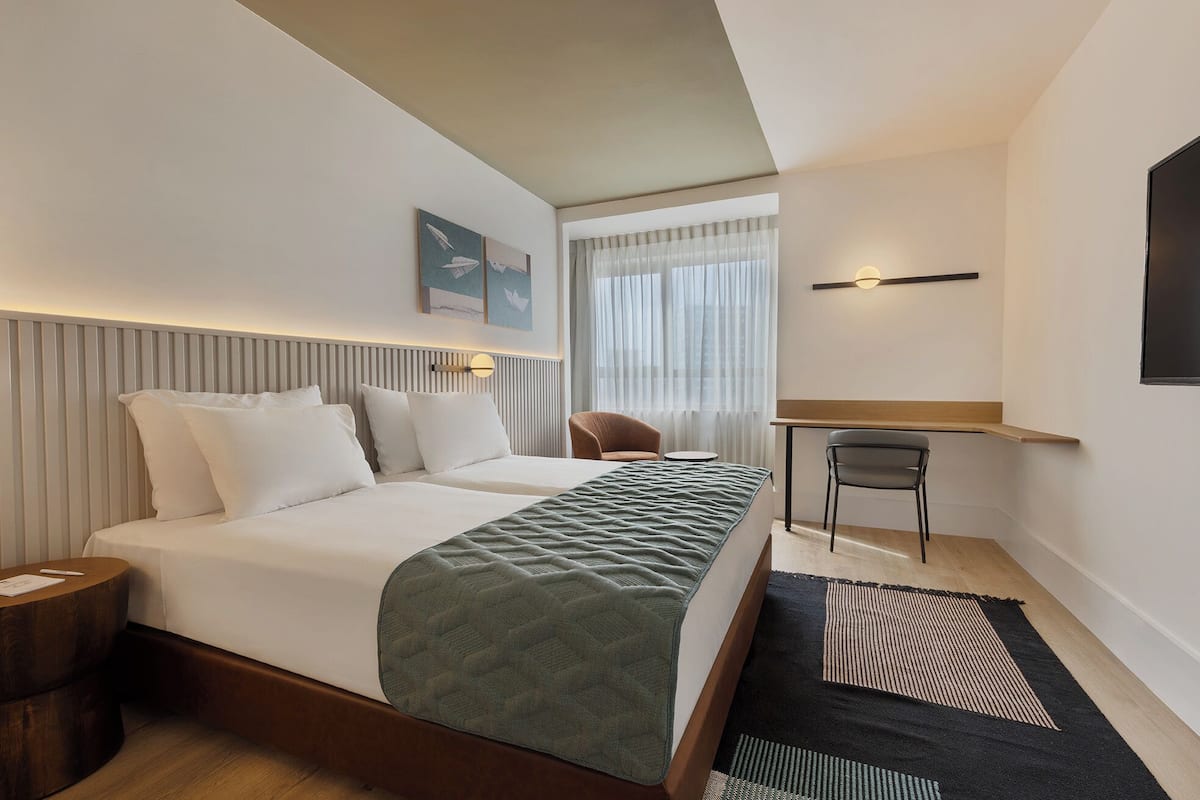 A hotel room with two beds, a desk, a chair, and a view of the city through sheer curtains.