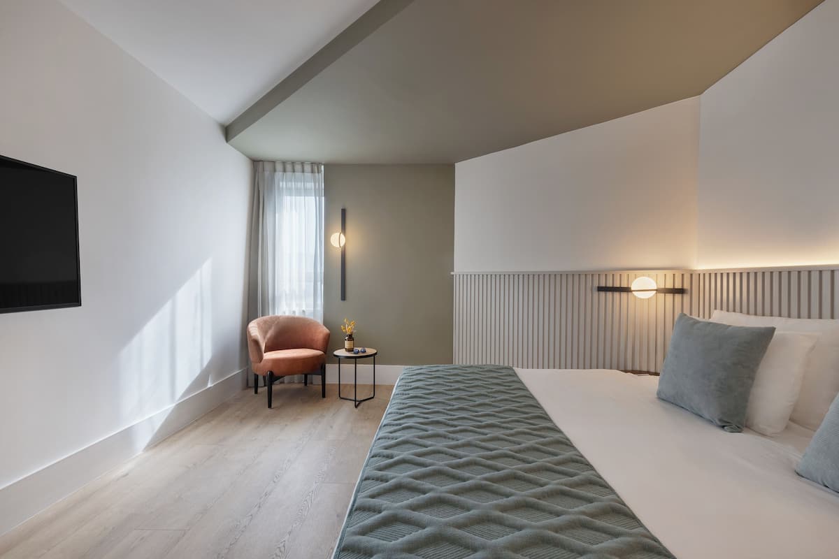A modern hotel room with a bed, a chair, a small table with a lamp, and a wall-mounted TV.