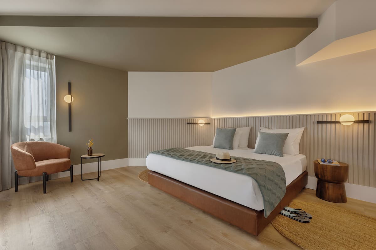 A modern hotel room with a large bed, a sitting area with a chair, a small table, and a nightstand.