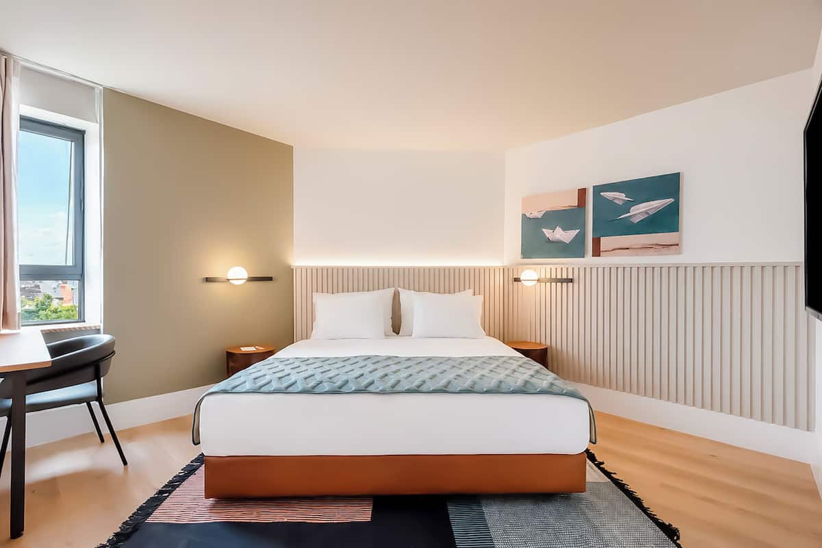 A modern hotel room with a bed, bedside tables, a desk, and a window.
