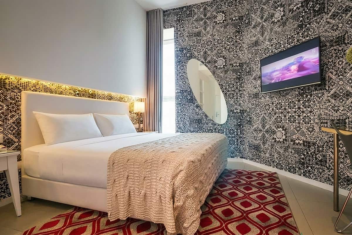 A modern hotel room with a bed, a TV, a mirror, and a patterned wall.