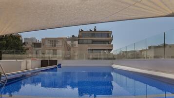 A rooftop swimming pool with clear blue water, surrounded by modern buildings and a glass barrier.
