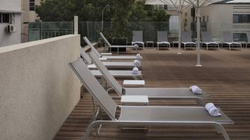 A rooftop lounge area with sun loungers and umbrellas, surrounded by a wooden deck and a glass fence.