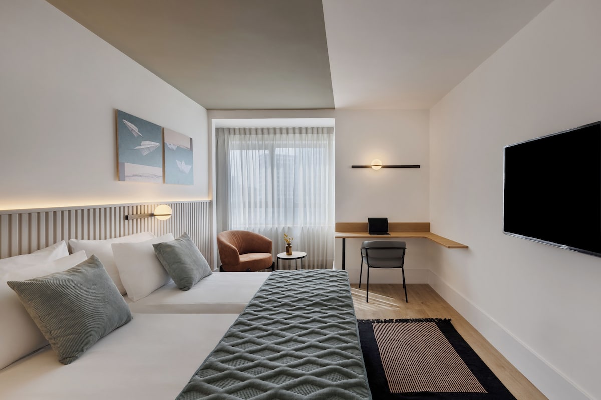 A modern hotel room with a bed, a desk, a chair, a TV, and artwork on the wall.
