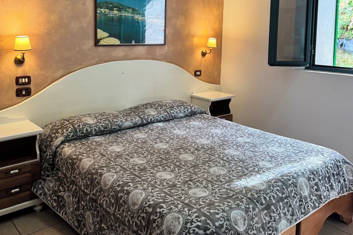 A bedroom with a bed, two bedside tables, a nightstand, a wall-mounted picture, and a window with a view of greenery.
