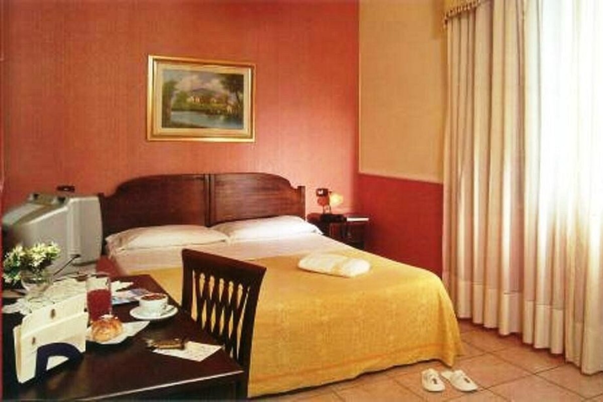 A hotel room with a bed, a desk with a chair, a lamp, and a painting on the wall.