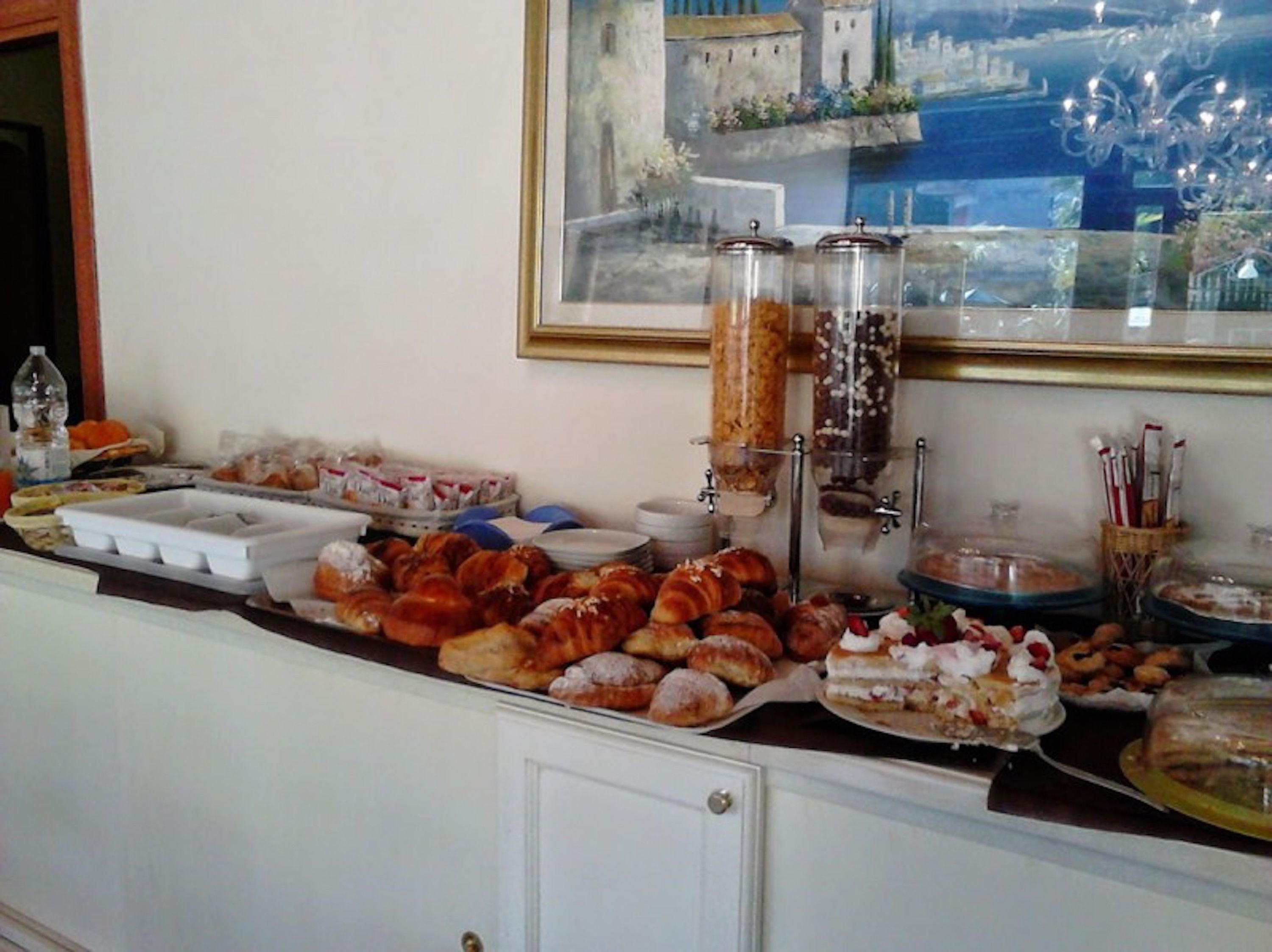 A buffet with various pastries and desserts, including croissants and cakes, displayed on a counter with a decorative mirror and a view of a coastal scene.