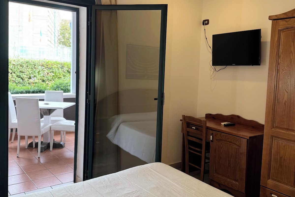 A bedroom with a bed, a wooden wardrobe, a TV mounted on the wall, and a sliding glass door leading to an outdoor seating area with chairs and a table.