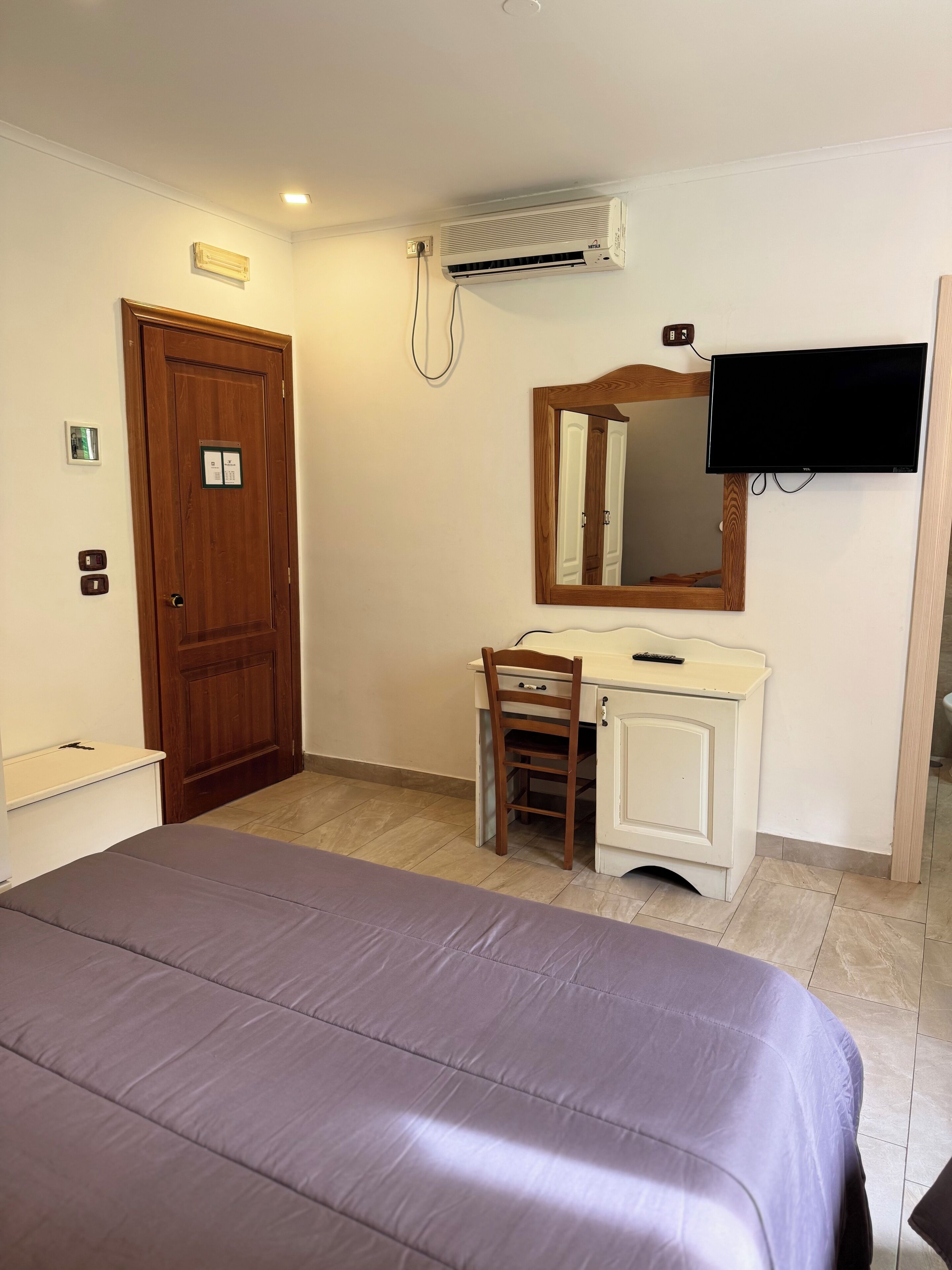 A bedroom with a bed, a wooden desk with a chair, a mirror, a television, and an air conditioning unit.