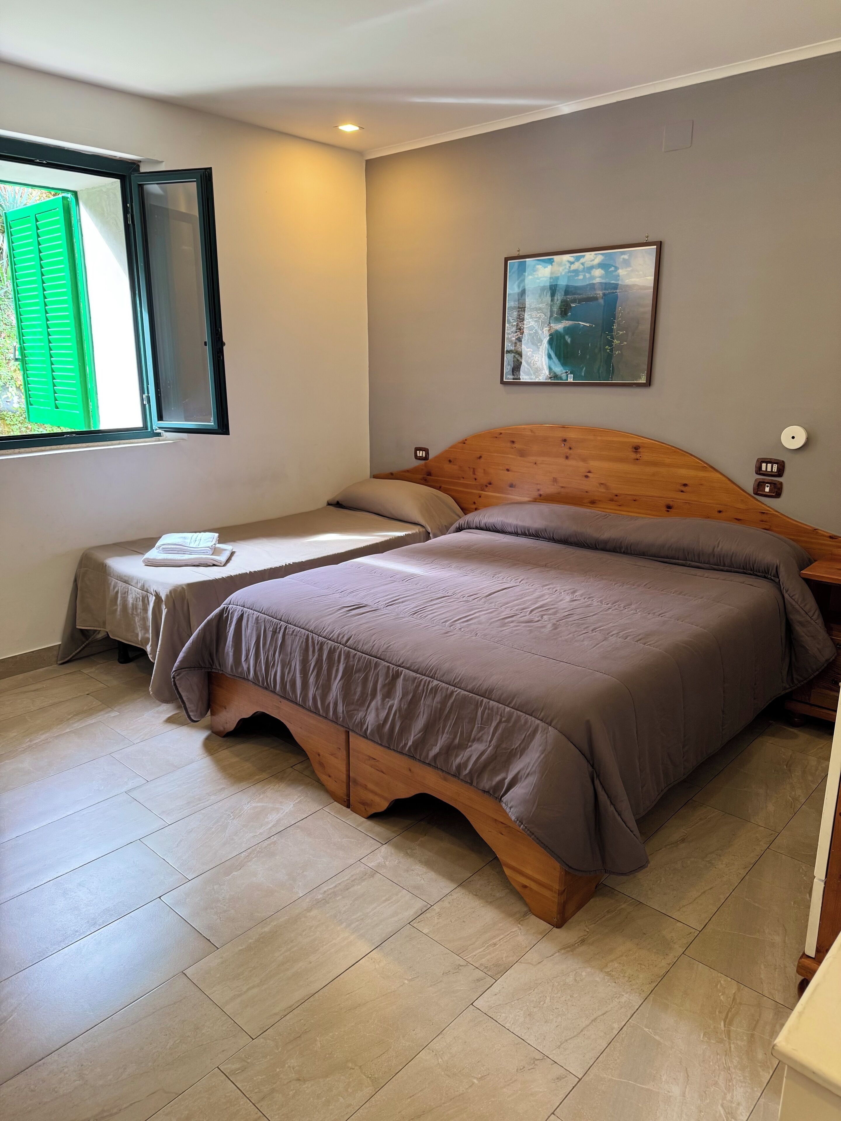 A bedroom with a wooden bed, a small bedside table, a nightstand, a window with green shutters, and a painting on the wall.