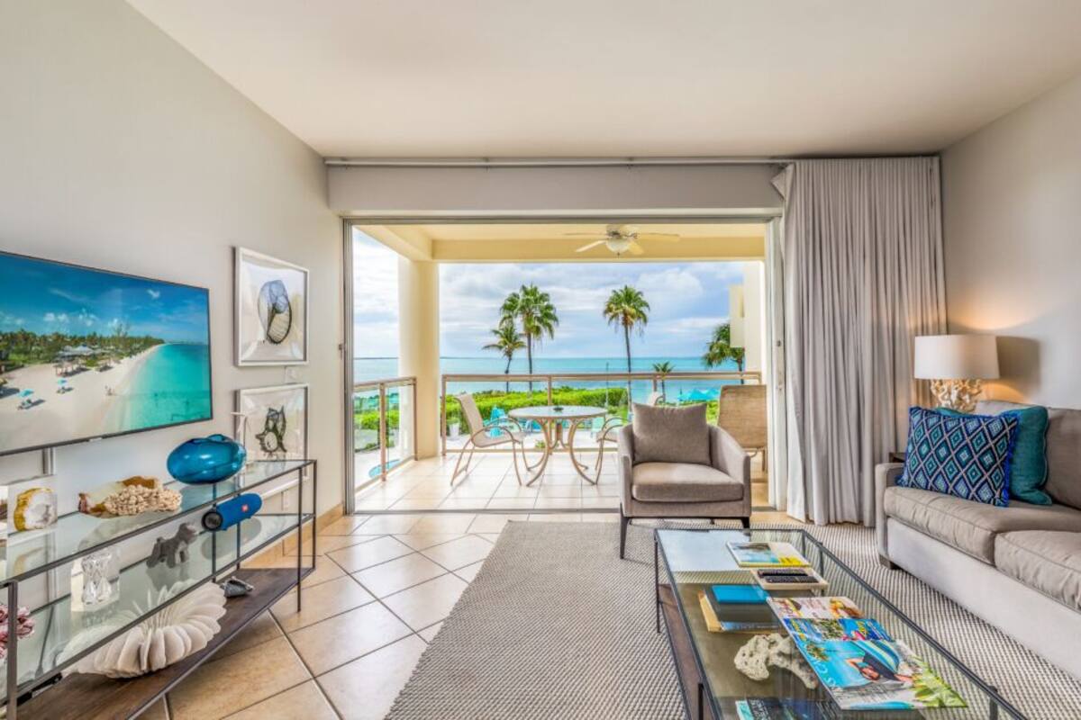 A modern living room with a large window overlooking a pool and palm trees, a sofa, a coffee table with magazines, and a glass table with decorative items.